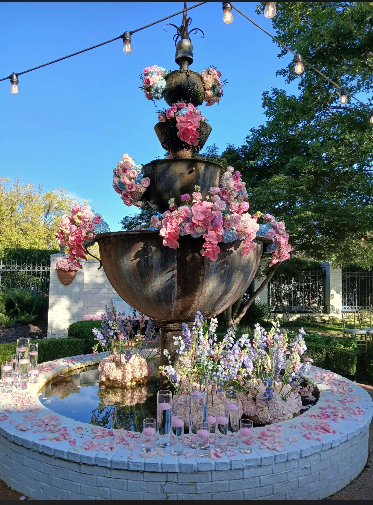 Tiered bronze fountain decorated with pink and white floral arrangements, surrounded by a white brick basin with candles and flower stems, beneath strung Edison bulbs in a garden setting. Bloomin’ Hills Flowers — Tillatoba, Mississippi.