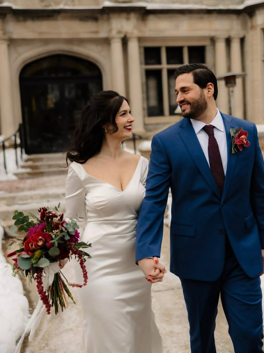 A bride in a white off-shoulder gown and a groom in a blue suit holding hands outside a historic stone building, the bride carrying a bouquet of deep red roses and burgundy amaranthus. Wild Child Fabrications Hair & Makeup — Atlanta, In town & north metro.