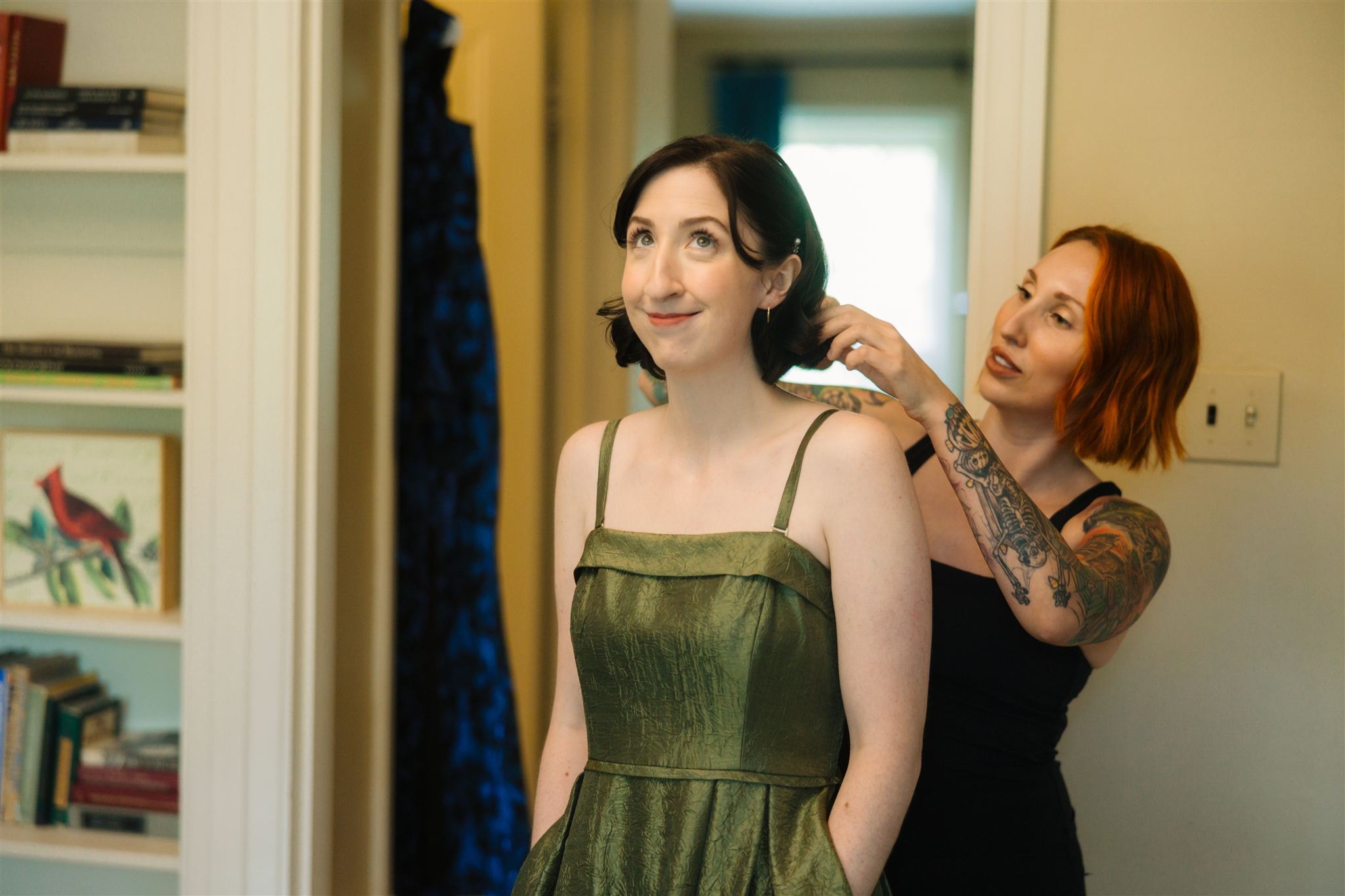 A woman in an olive green dress stands facing left while another woman with red hair and visible arm tattoos adjusts her hair from behind in a home interior with bookshelves. Wild Child Fabrications Hair & Makeup — Atlanta, In town & north metro.