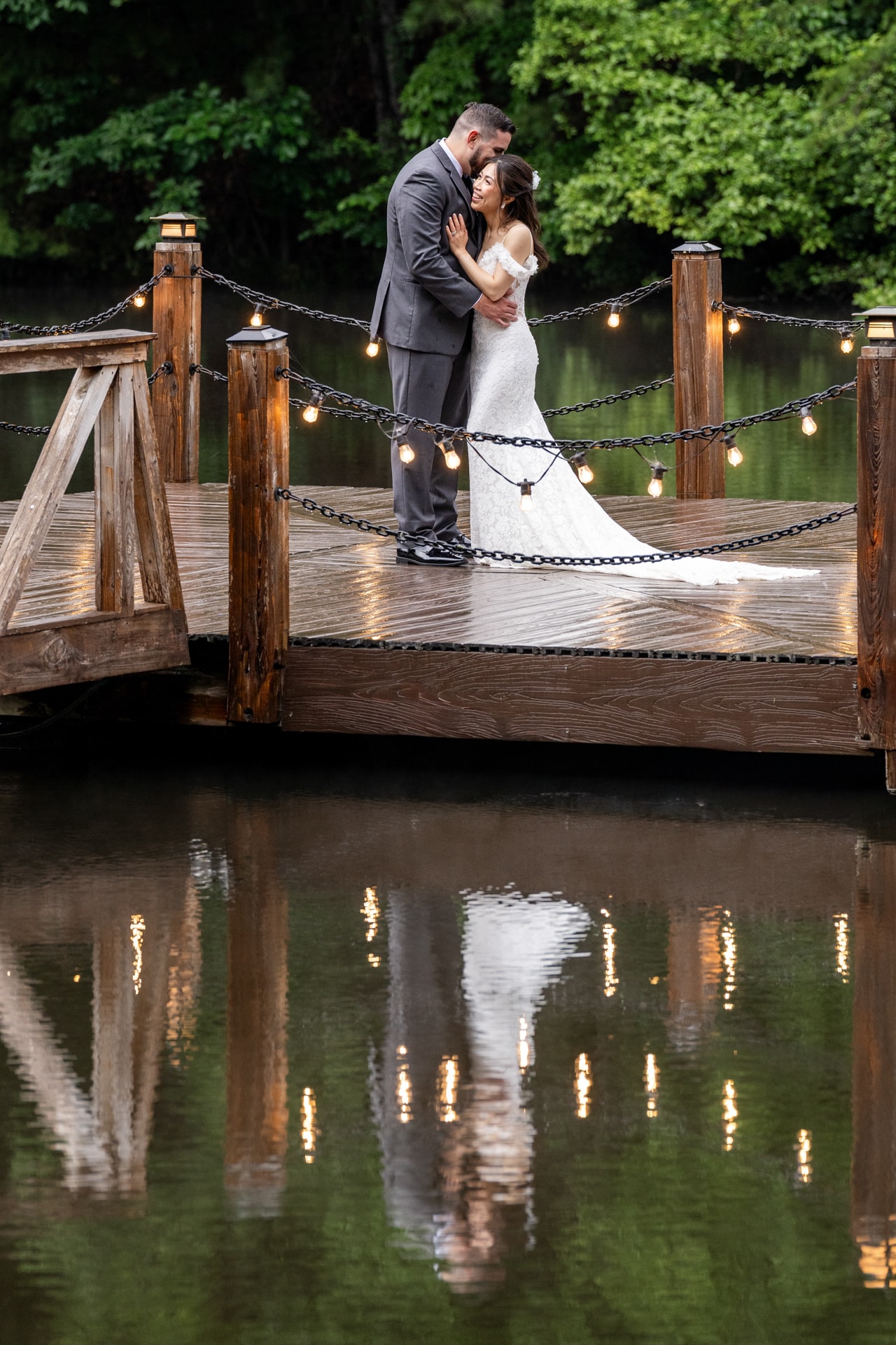 A groom in a gray suit and bride in a white wedding dress embrace on a wooden dock over still water, with string lights and black chain railings framing the scene and green foliage in the background. Double N Media — Atlanta, Southeast.