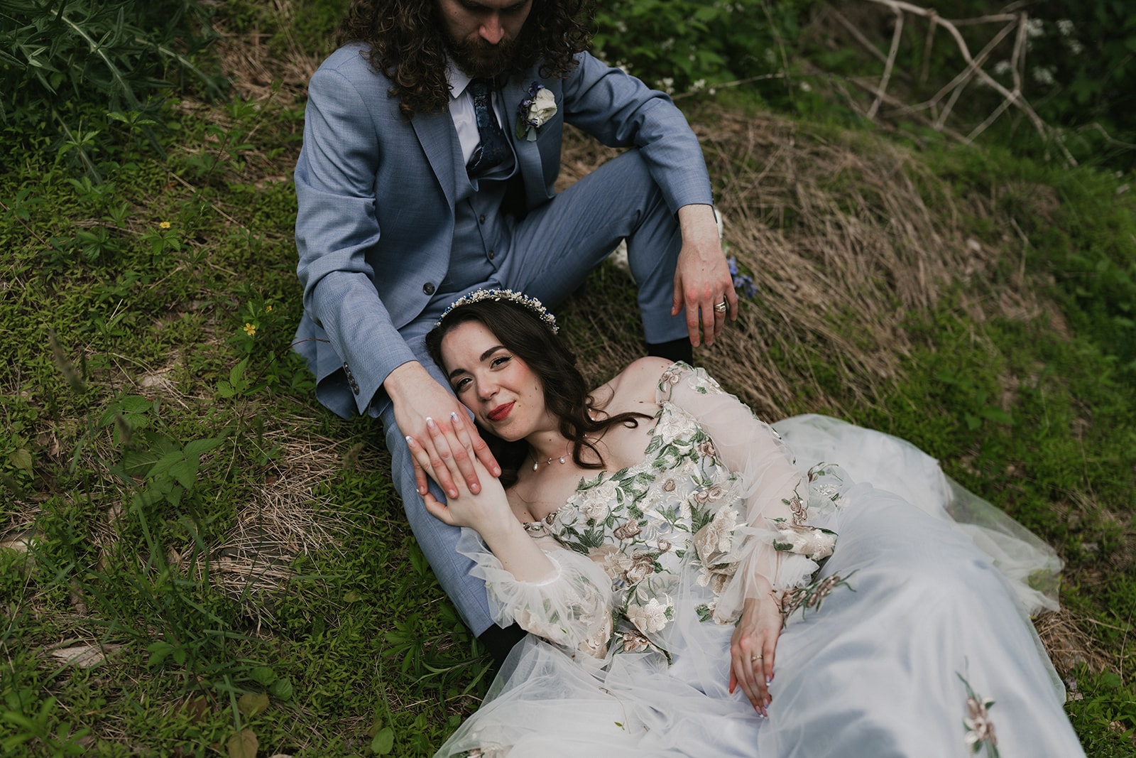A bride and groom recline together on grass and moss, the bride wearing an embroidered gown with tulle skirt and floral crown, the groom in a slate blue suit with boutonniere, surrounded by overgrown vegetation and branches. Wild Child Fabrications Hair & Makeup — Atlanta, In town & north metro.