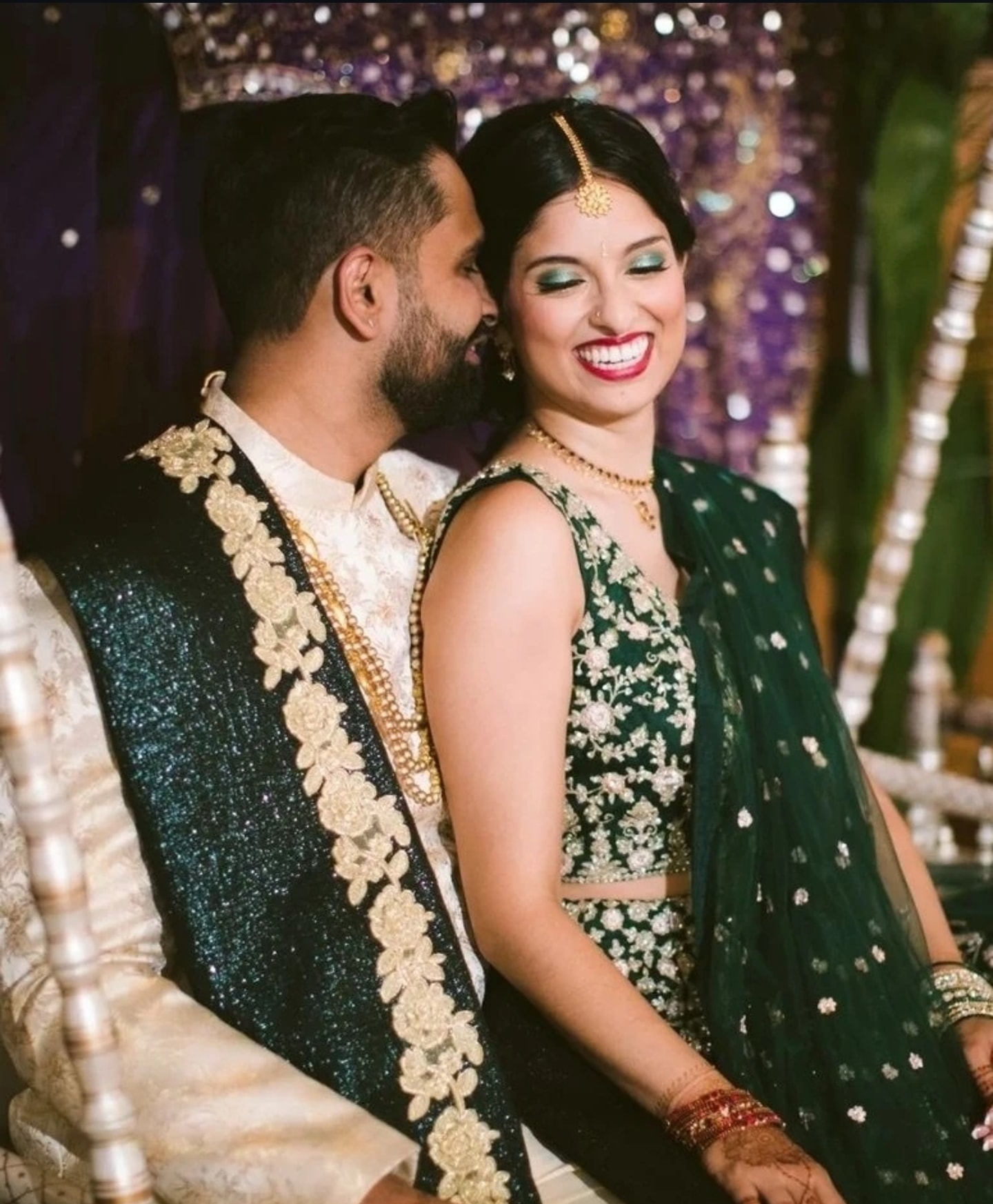 Groom in teal sequin sherwani with gold embroidery embracing bride in emerald lehenga with floral embroidery and gold jewelry, against a purple and gold bokeh backdrop. Wild Child Fabrications Hair & Makeup — Atlanta, In town & north metro.