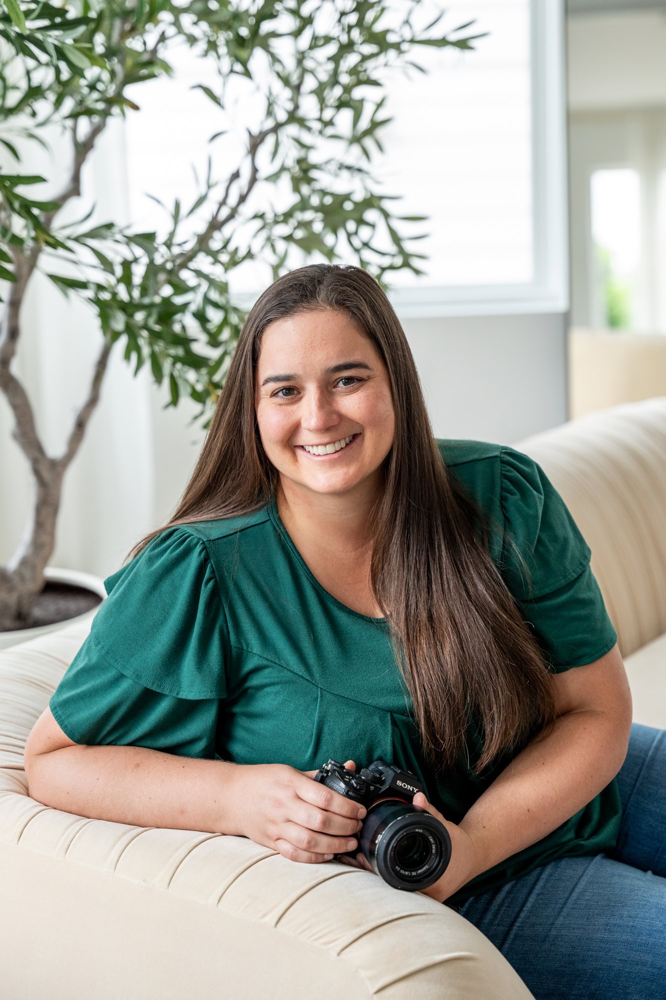 Woman in a dark green short-sleeved top sitting on a cream sofa, holding a camera, with an olive tree in a white pot visible behind her. Double N Media — Atlanta, Southeast.