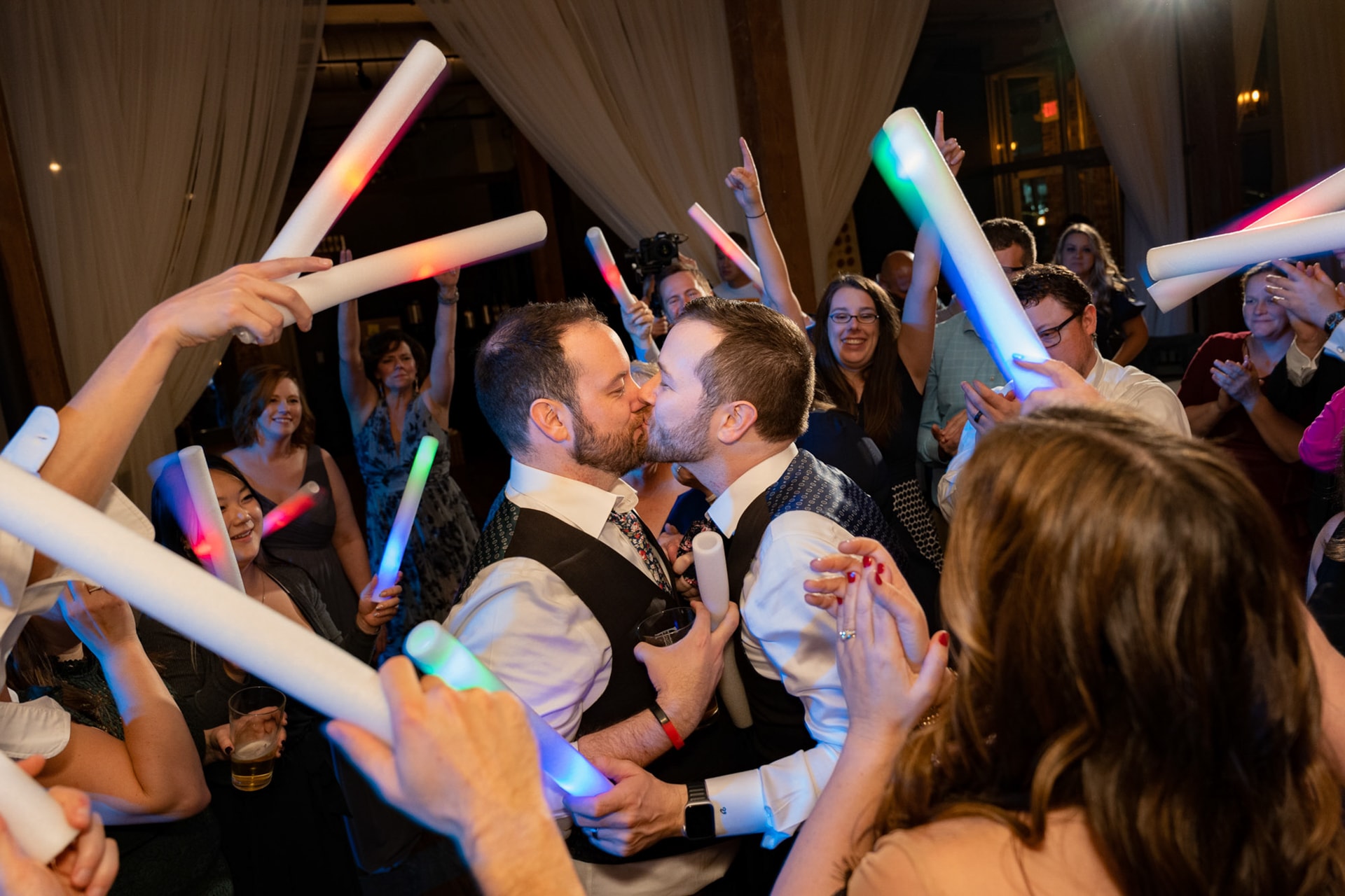 Two grooms kiss at the center of a dance floor surrounded by guests holding illuminated foam light-up batons in multicolored light, with draped fabric walls and a historic ballroom interior visible behind them. Double N Media — Atlanta, Southeast.