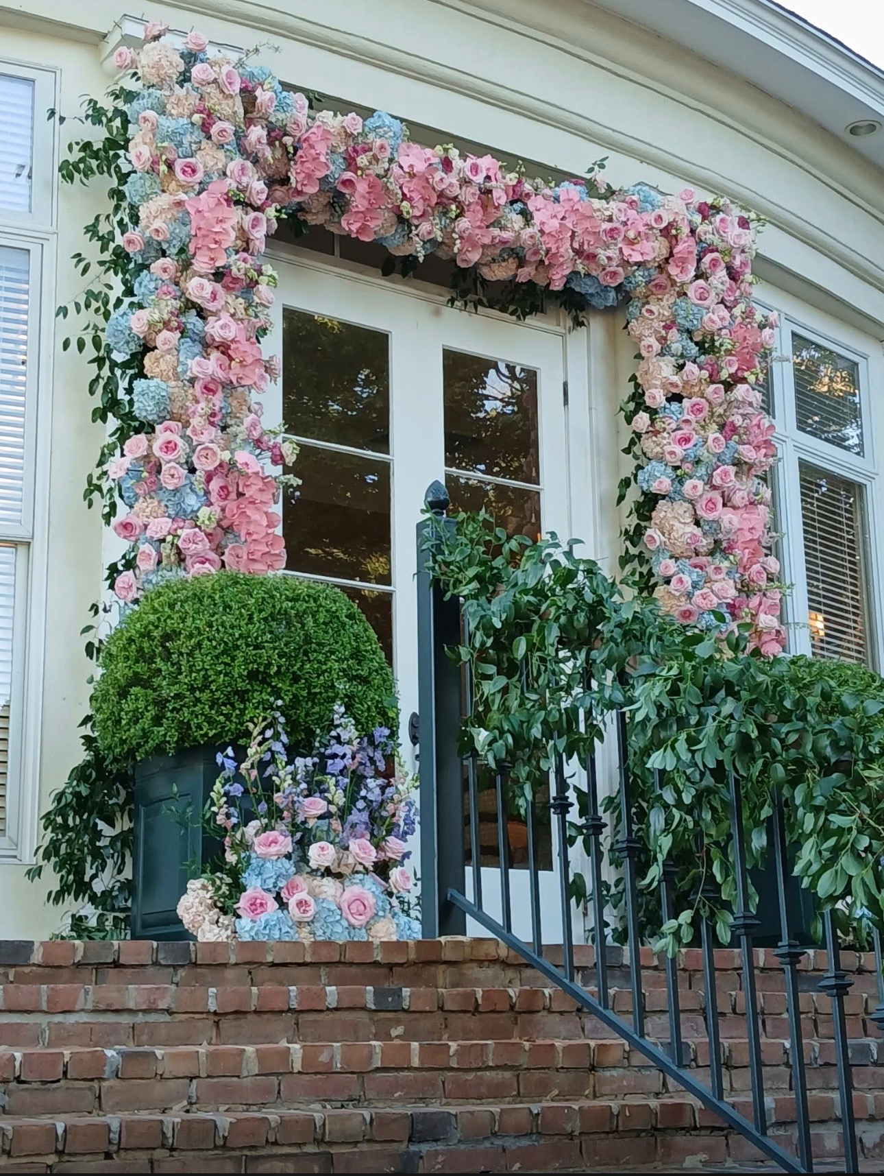 Floral arch installation framing a black door and windows with pink roses, blue hydrangeas, cream peonies, green foliage, and a boxwood topiary at the entrance of a white brick building. Bloomin’ Hills Flowers — Tillatoba, Mississippi.