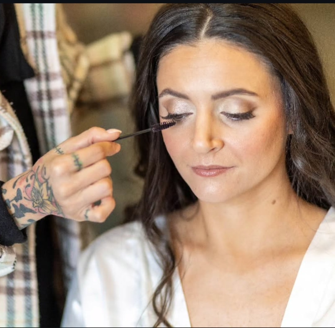 A makeup artist with tattooed hands applies mascara to a woman with dark hair and neutral makeup during a bridal beauty preparation. Wild Child Fabrications Hair & Makeup — Atlanta, In town & north metro.