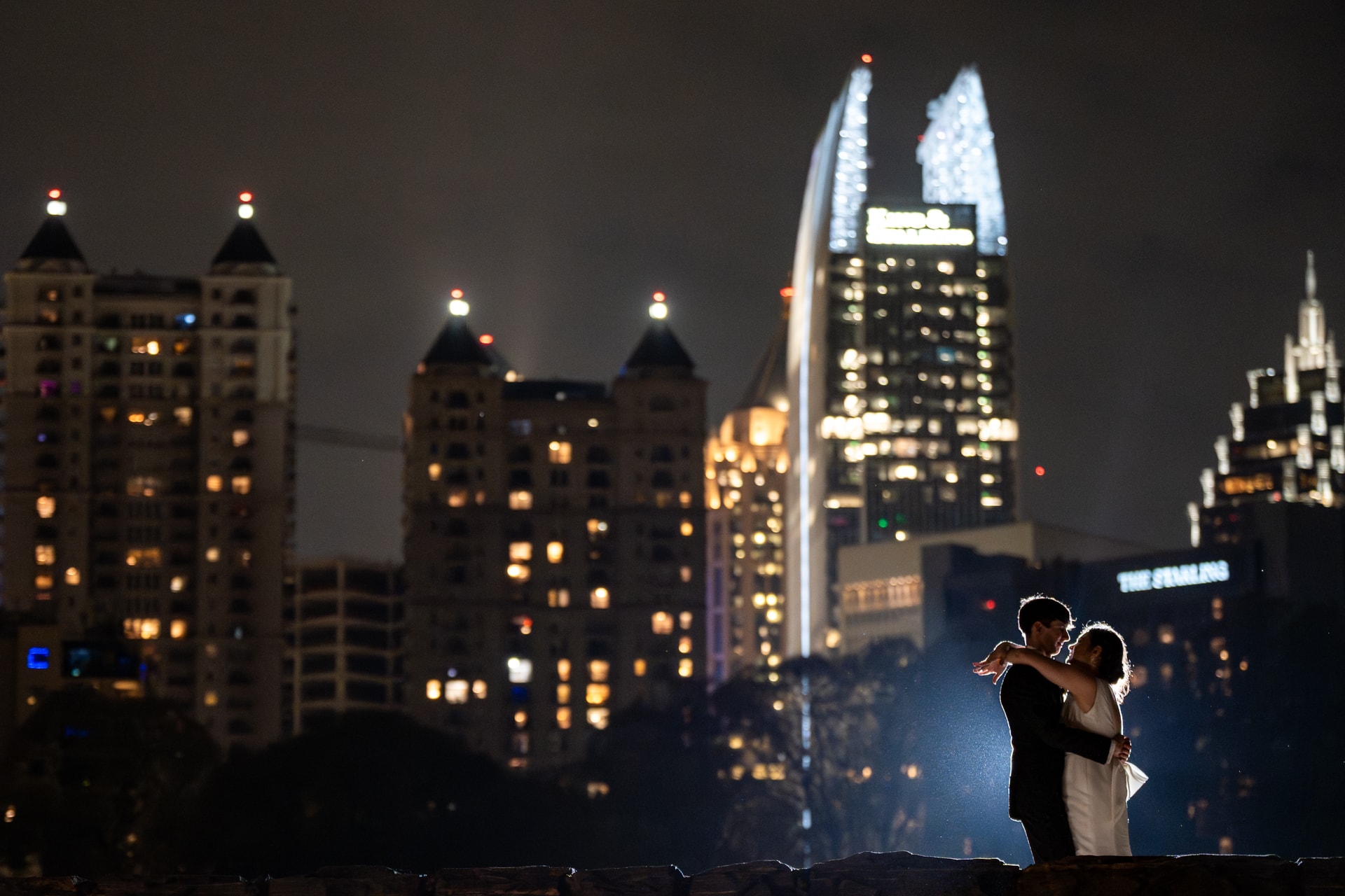 A groom in a dark suit and bride in a white wedding dress embrace on a rooftop at night, with an illuminated city skyline and lit skyscrapers in the background. Double N Media — Atlanta, Southeast.