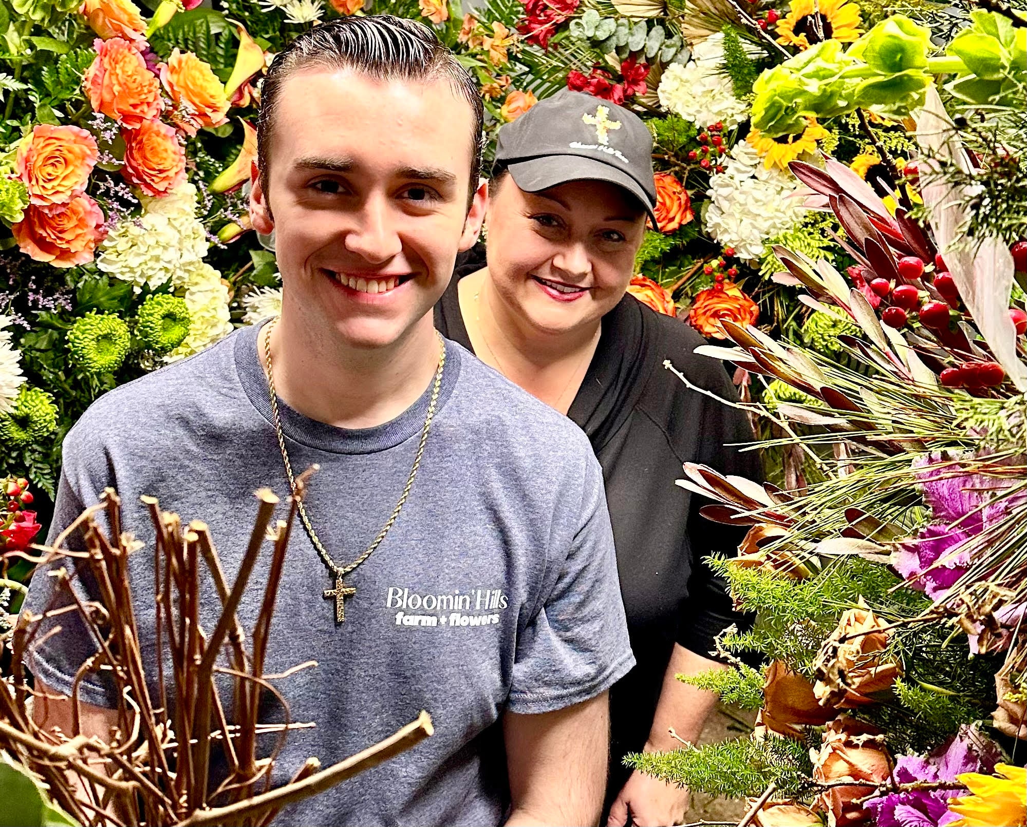Two people standing in front of a dense flower wall of multicolored roses, hydrangeas, dahlias, and greenery. The person in front wears a gray t-shirt with text and a gold chain necklace, smiling at the camera. Bloomin’ Hills Flowers — Tillatoba, Mississippi.