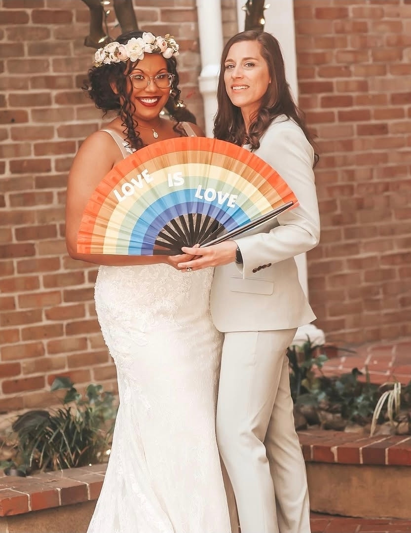 Two brides in white wedding attire hold a rainbow fan printed with "Love is Love" against a red brick wall backdrop. Wild Child Fabrications Hair & Makeup — Atlanta, In town & north metro.