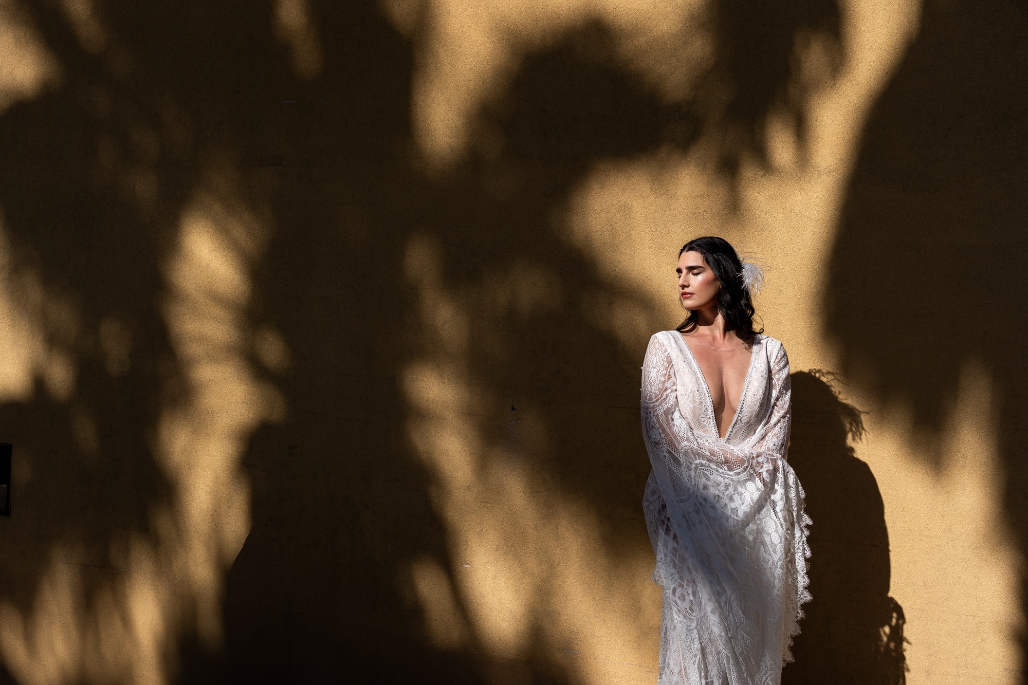 Woman in a white lace wrap dress standing against a tan stucco wall with dappled tree shadows. Double N Media — Atlanta, Southeast.