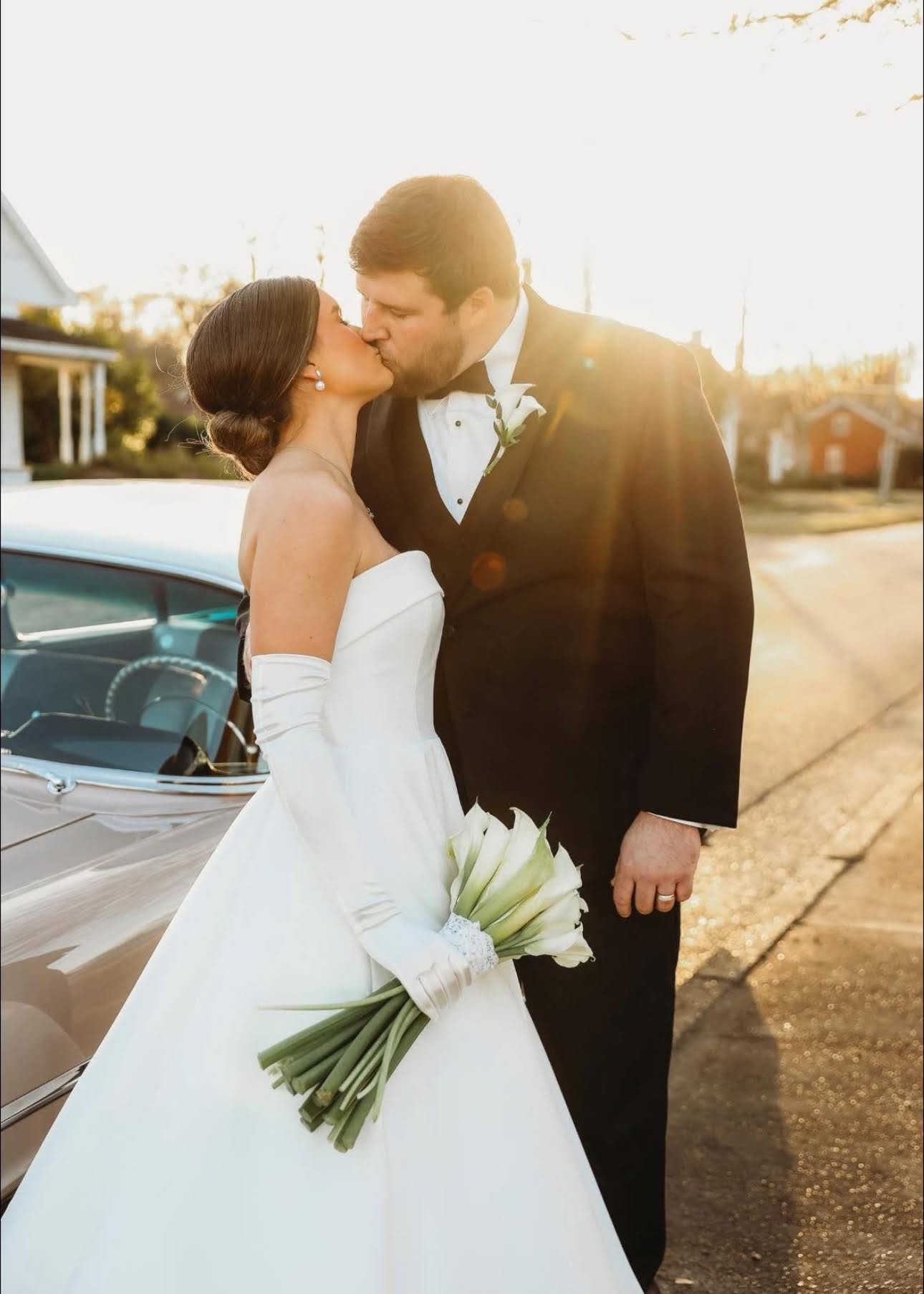 A bride in a white strapless gown and groom in a black tuxedo kiss beside a convertible car at sunset, the bride holding a bouquet of white calla lilies with green stems wrapped in white ribbon. Bloomin’ Hills Flowers — Tillatoba, Mississippi.