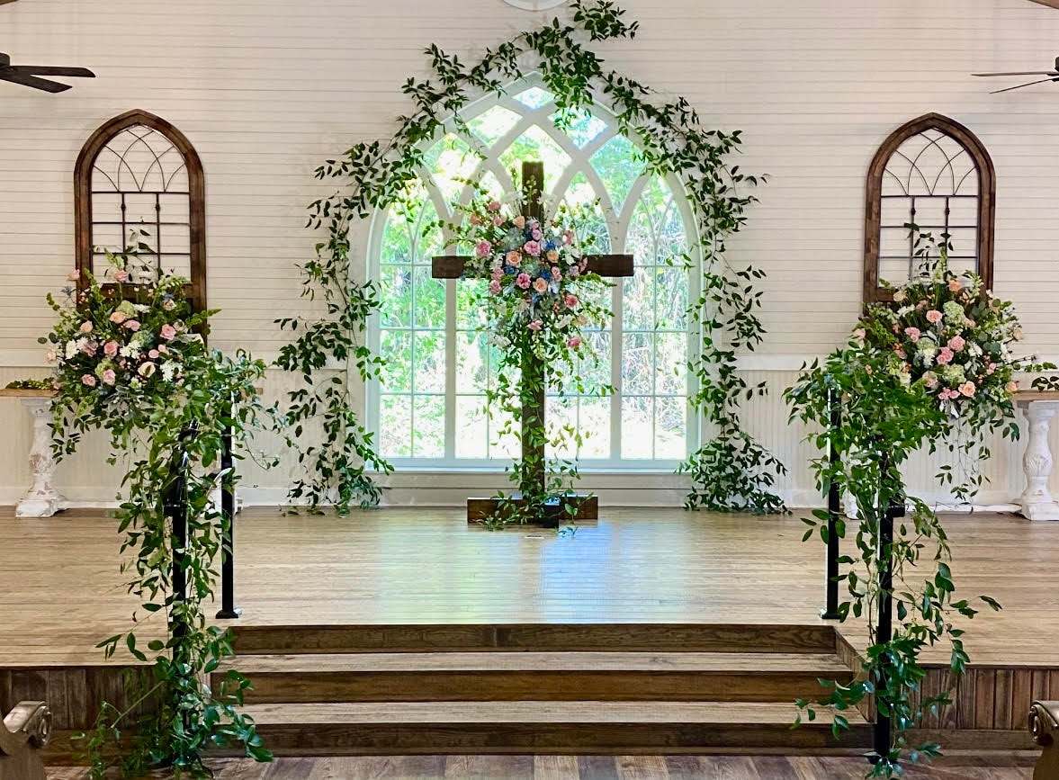 A church altar adorned with a wooden cross draped in cascading greenery and pink flowers, flanked by two black metal arbors wrapped in ivy and trailing vines, with arched windows and pink floral arrangements on pedestals on either side. Bloomin’ Hills Flowers — Tillatoba, Mississippi.