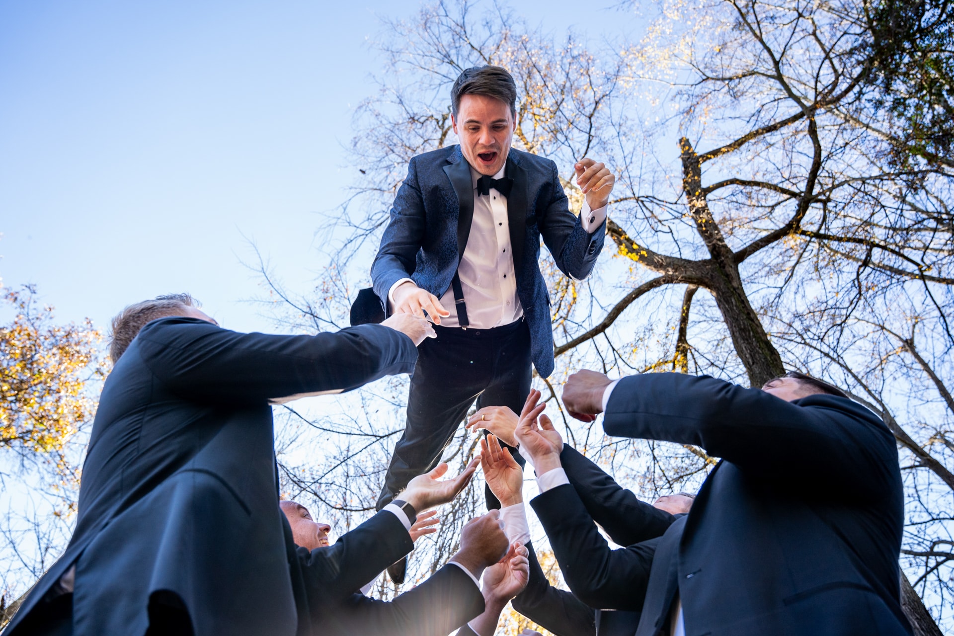 Groom in navy blazer and bow tie lifted on shoulders by groomsmen in dark suits beneath bare trees against blue sky. Double N Media — Atlanta, Southeast.