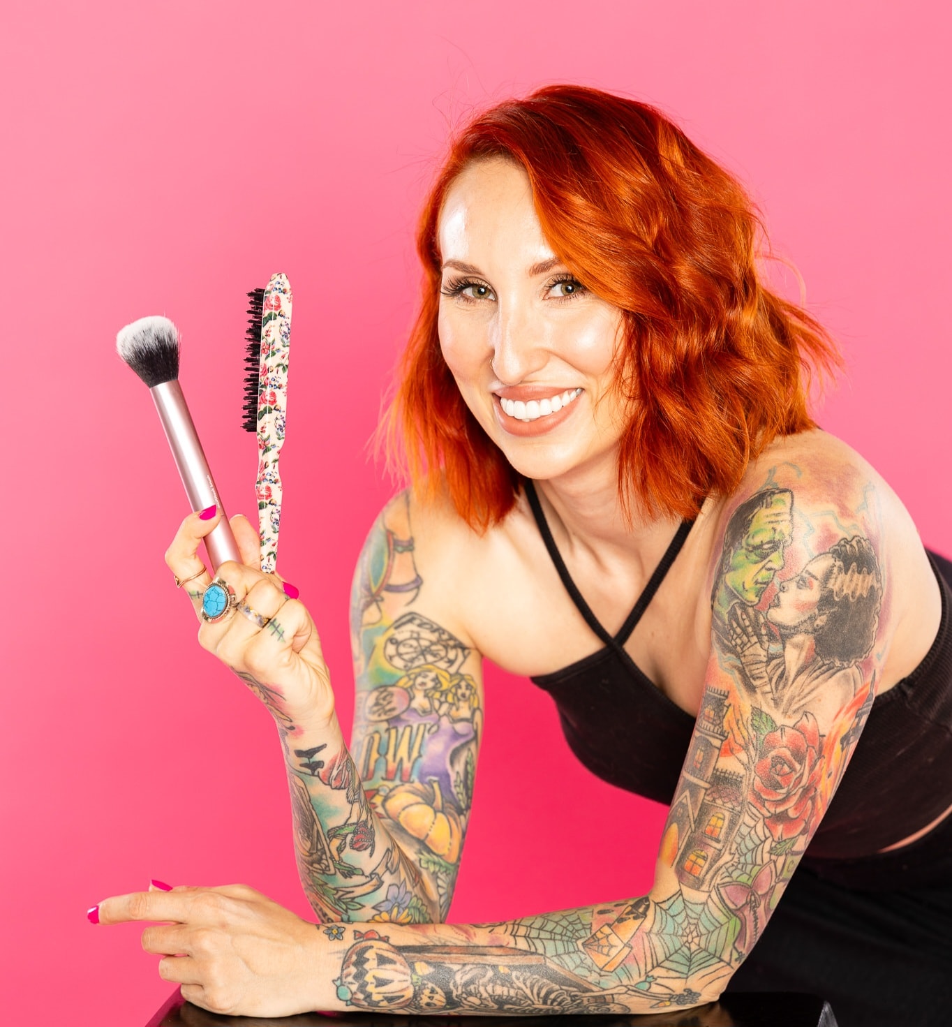 Woman with orange wavy hair and full-sleeve tattoos holding a makeup brush and floral-patterned hair brush against a pink background. Wild Child Fabrications Hair & Makeup — Atlanta, In town & north metro.