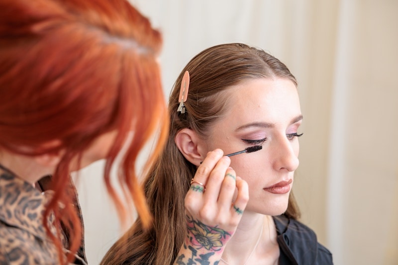 A makeup artist with red hair and visible tattoos applies mascara to a client with brown hair and a peach hair clip. Wild Child Fabrications Hair & Makeup — Atlanta, In town & north metro.
