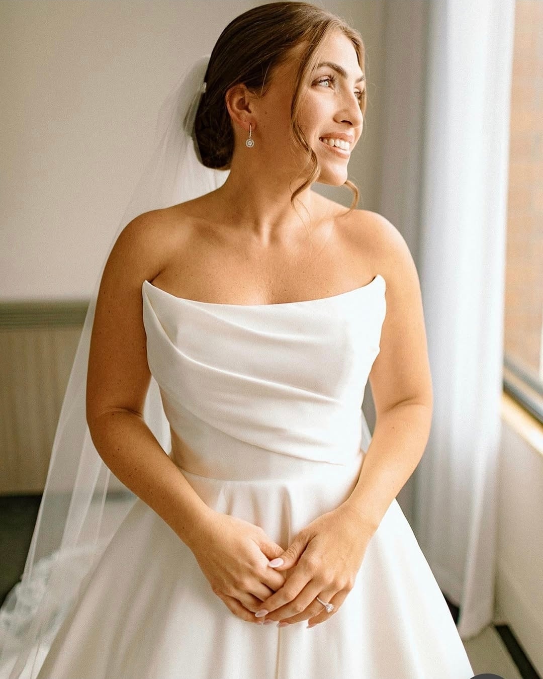 Woman wearing a white strapless wedding dress with cowl neckline and veil, smiling and looking to the side in a bright interior space. Wild Child Fabrications Hair & Makeup — Atlanta, In town & north metro.