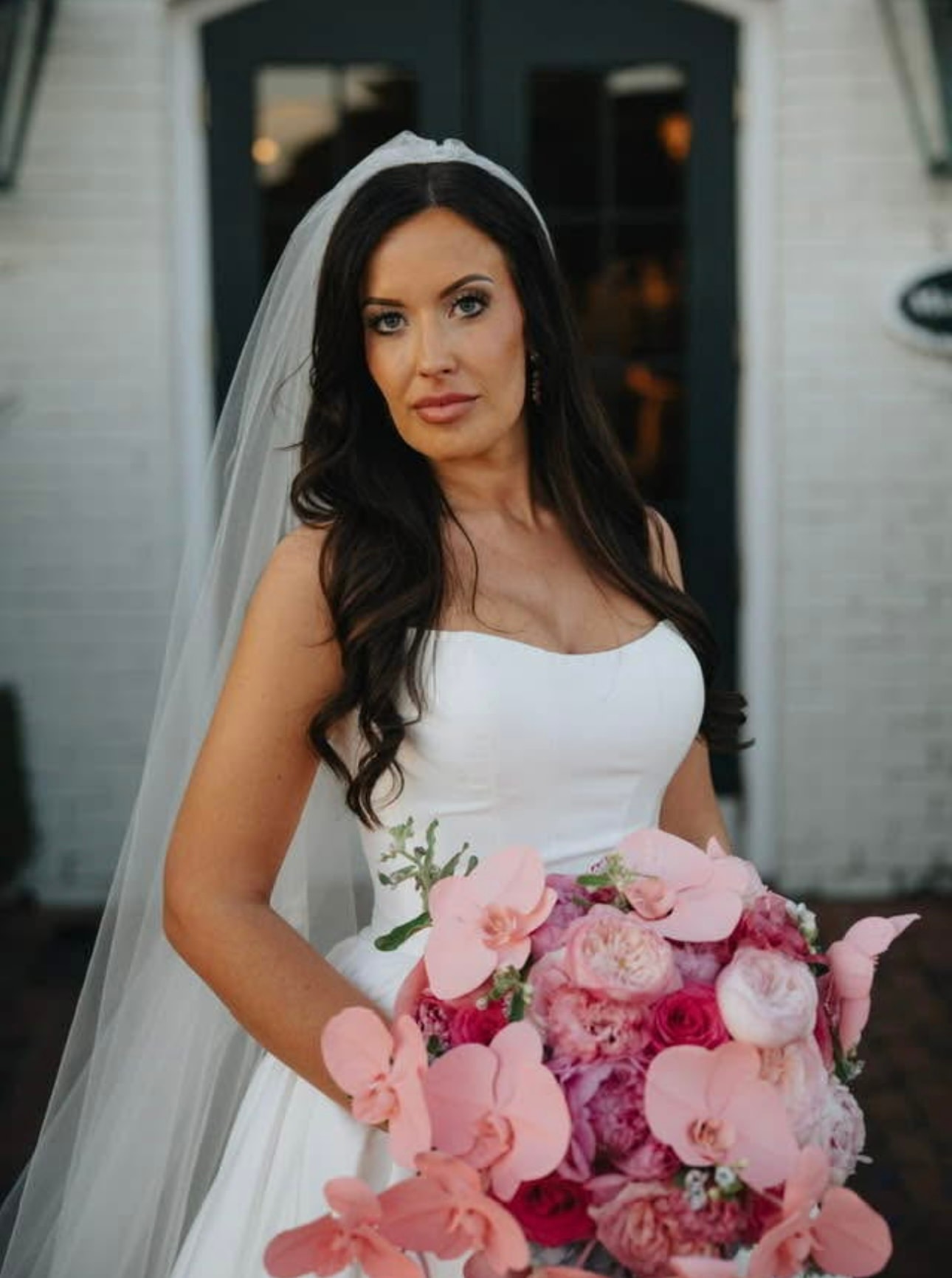 Bride in white sleeveless gown and sheer veil holding bridal bouquet of pink orchids, garden roses, and peonies in front of a whitewashed building with dark doors. Bloomin’ Hills Flowers — Tillatoba, Mississippi.