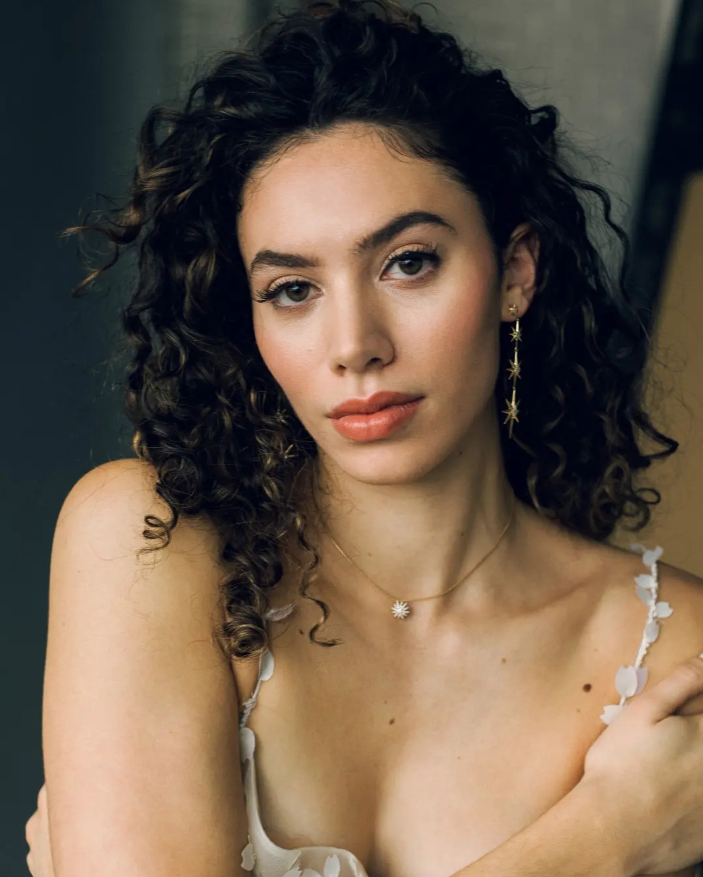Woman with dark curly hair wearing a white strappy top, gold star earrings, and a daisy pendant necklace, gazing to the side against a neutral background. Wild Child Fabrications Hair & Makeup — Atlanta, In town & north metro.