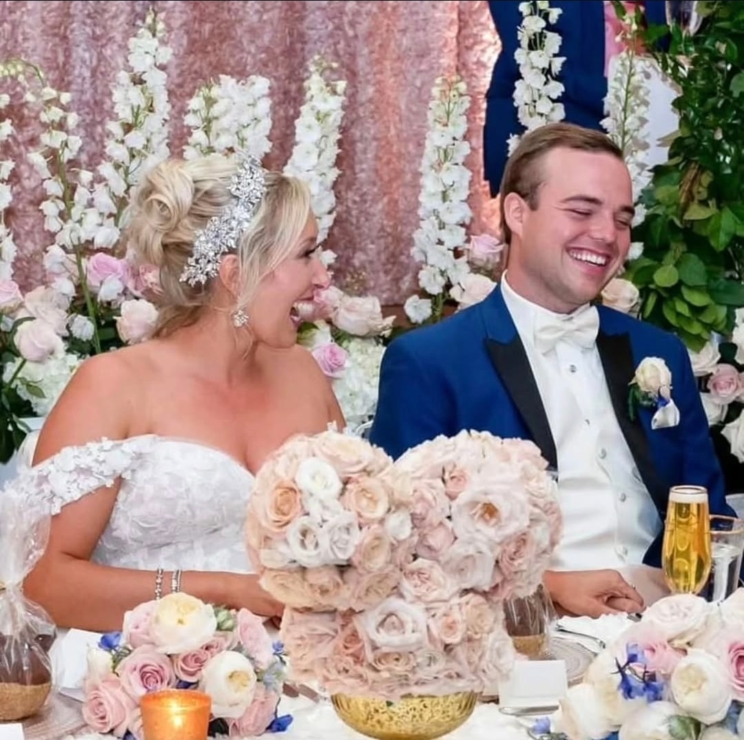Bride and groom laugh while seated at their reception table, surrounded by blush and white rose arrangements and tall white delphinium sprays against a pink sequined backdrop. Wild Child Fabrications Hair & Makeup — Atlanta, In town & north metro.