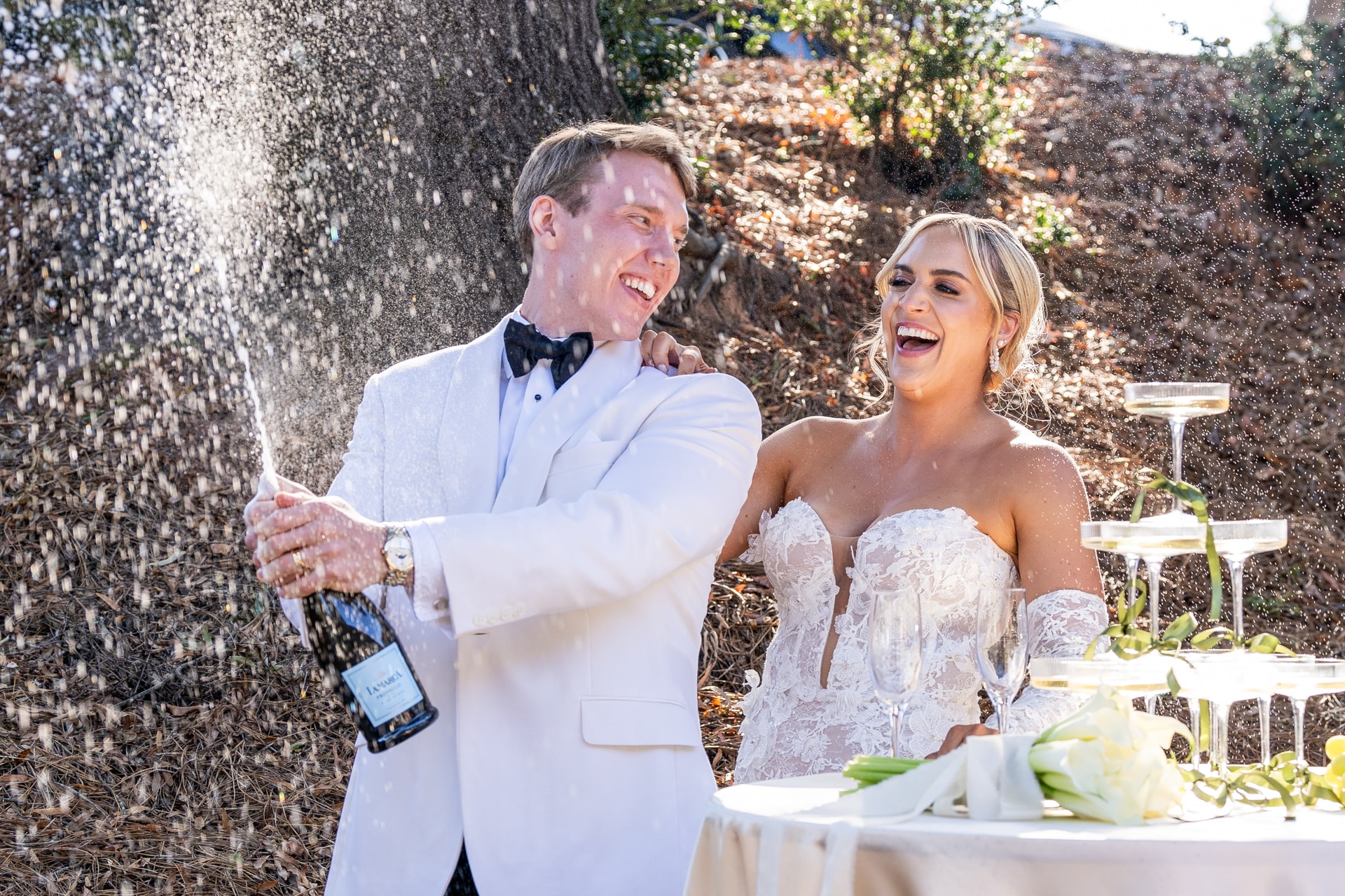 A groom in a white tuxedo jacket and bow tie sprays champagne while laughing beside a bride in a beaded strapless white gown at an outdoor garden reception near a stone wall. Double N Media — Atlanta, Southeast.