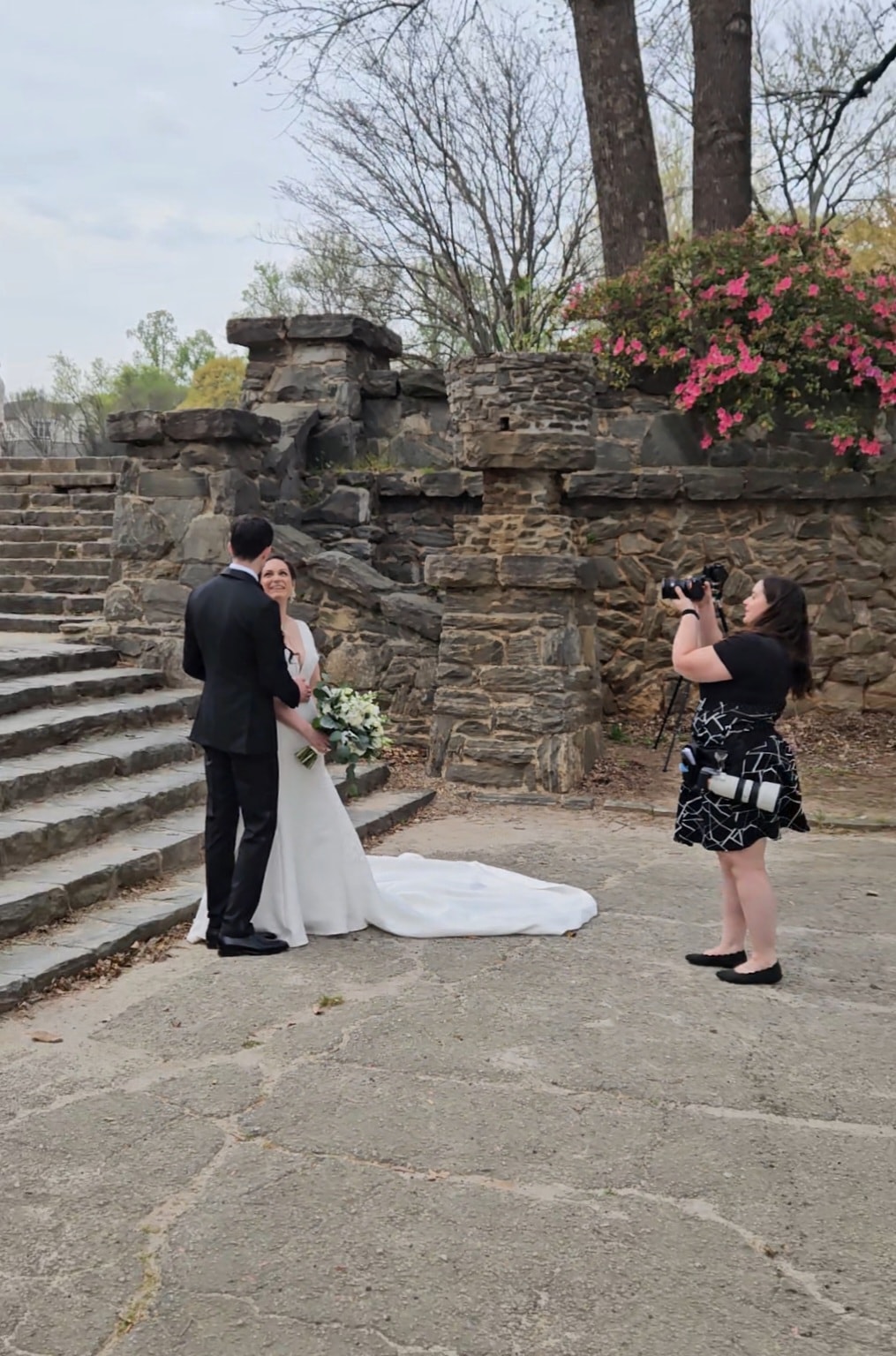 Photographer in black dress with camera photographs a bride and groom at a stone ruins venue with pink flowering shrubs and bare trees. Double N Media — Atlanta, Southeast.