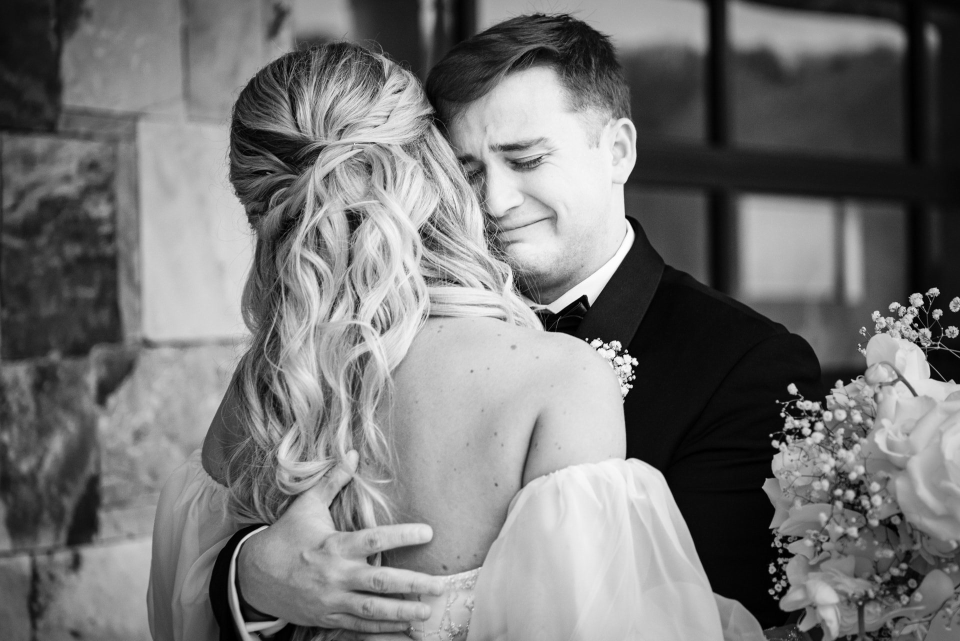 A bride with long wavy hair and a strapless white gown embraces a groom in a black tuxedo and bow tie, holding a bouquet of white roses and baby's breath in a stone courtyard. Double N Media — Atlanta, Southeast.