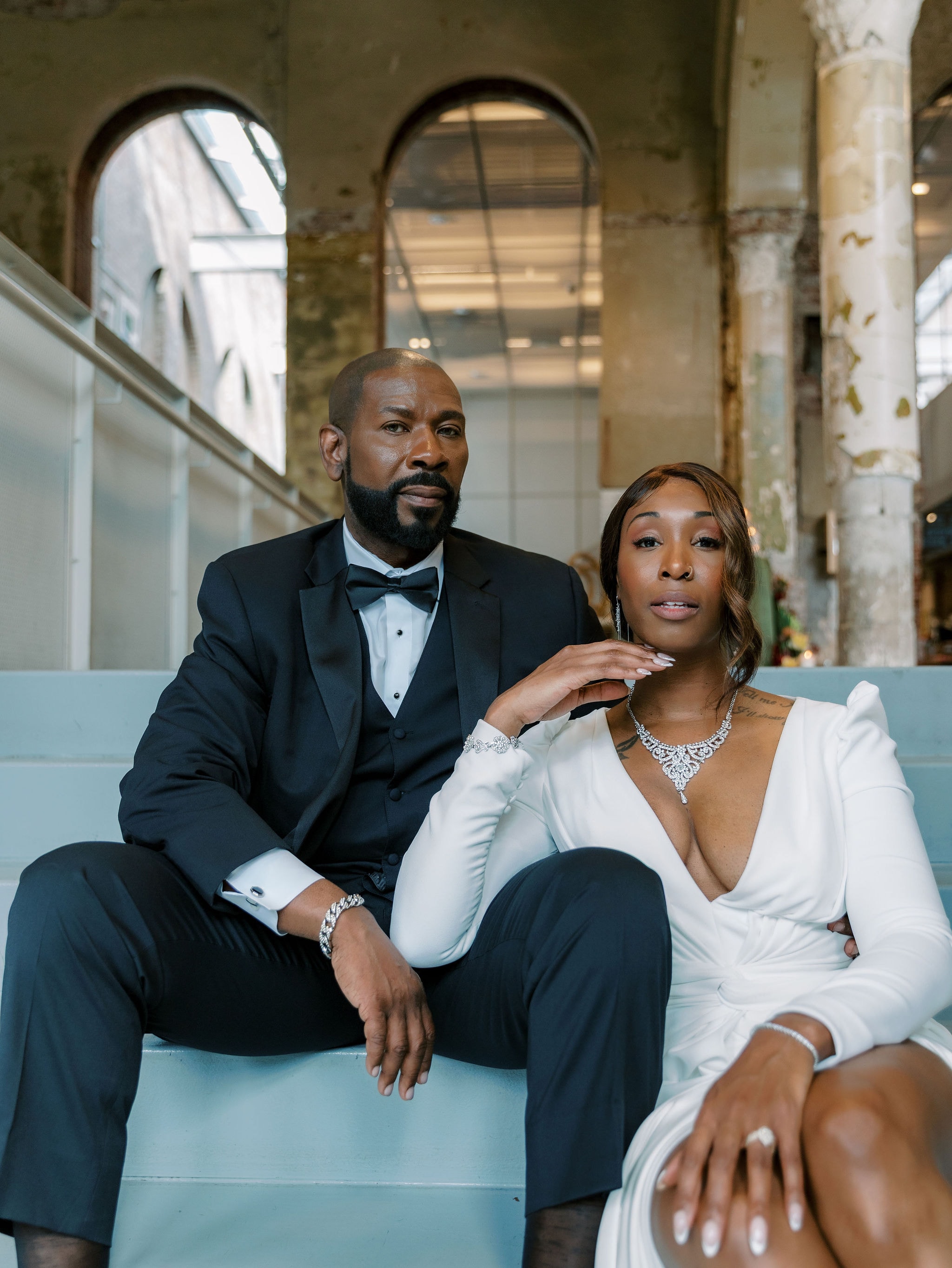 A man in a black tuxedo with bow tie and a woman in a white wrap blouse sit together on a light blue bench in an industrial loft space with exposed arches and columns. The woman wears a diamond necklace and bracelet. Wild Child Fabrications Hair & Makeup — Atlanta, In town & north metro.