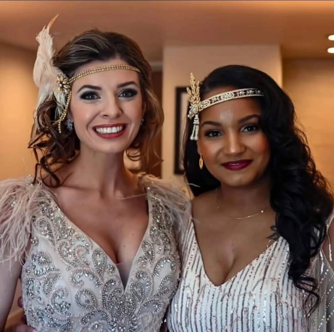 Two women wearing 1920s-inspired beaded dresses and jeweled headbands with feather and tassel accents, posing indoors in warm lighting. Wild Child Fabrications Hair & Makeup — Atlanta, In town & north metro.