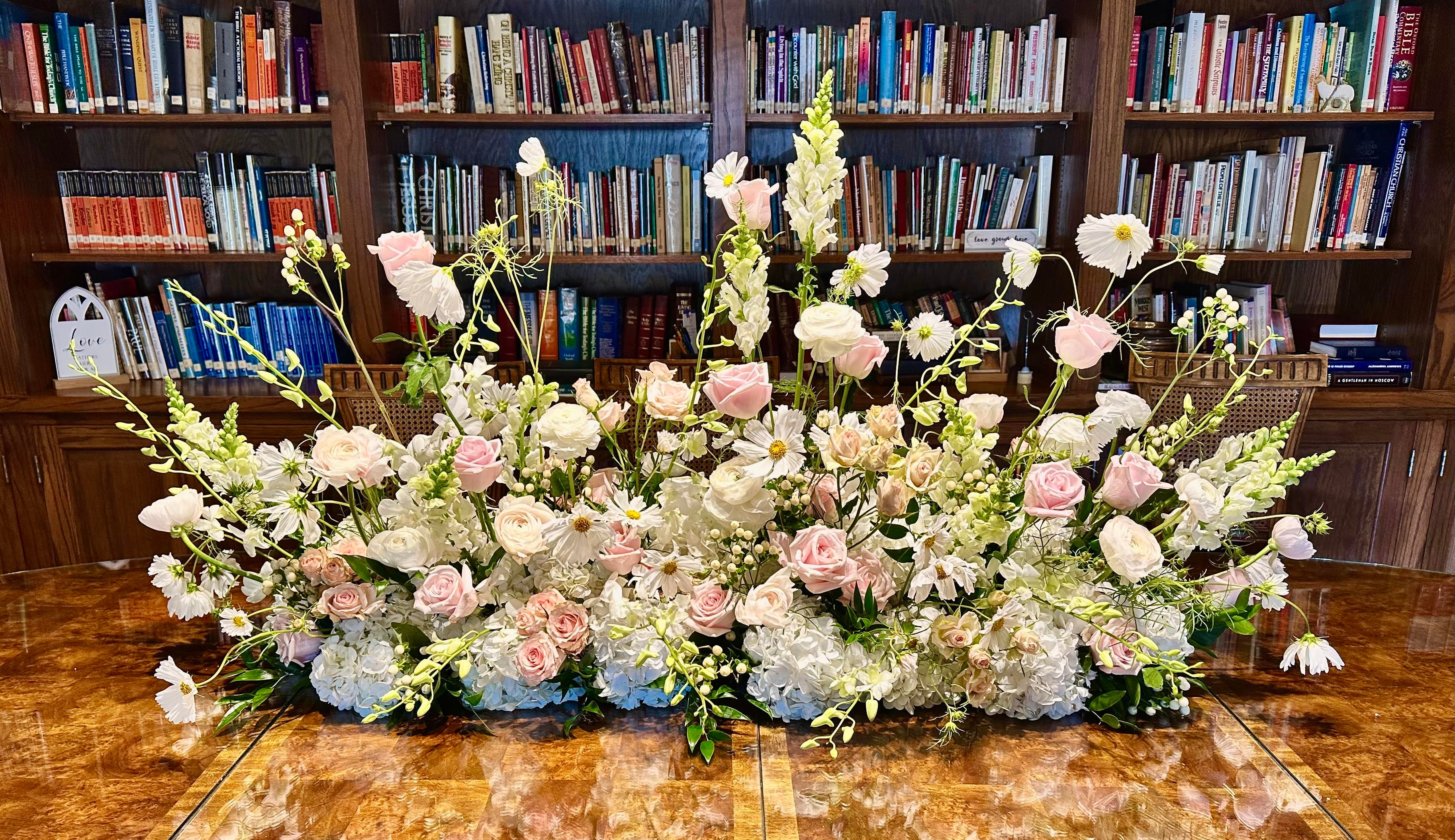 A low floral arrangement of pink and white roses, white hydrangeas, daisies, and green foliage sits on a polished wood floor in front of floor-to-ceiling wooden bookshelves in a library setting. Bloomin’ Hills Flowers — Tillatoba, Mississippi.