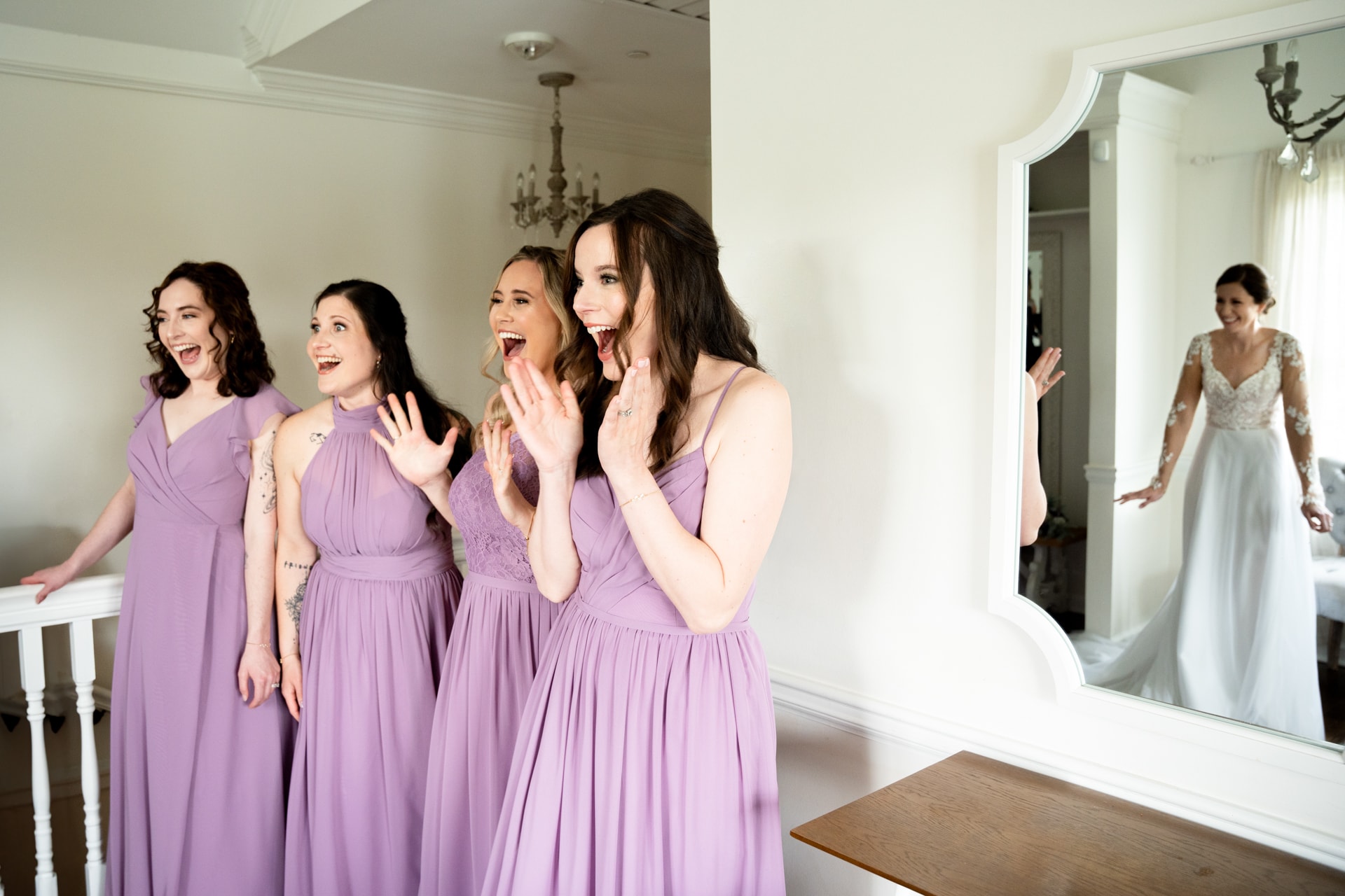 Four women in lavender bridesmaid dresses react with excitement while watching a bride in a beaded white gown enter through a mirror-lined dressing room in a bridal suite. Double N Media — Atlanta, Southeast.